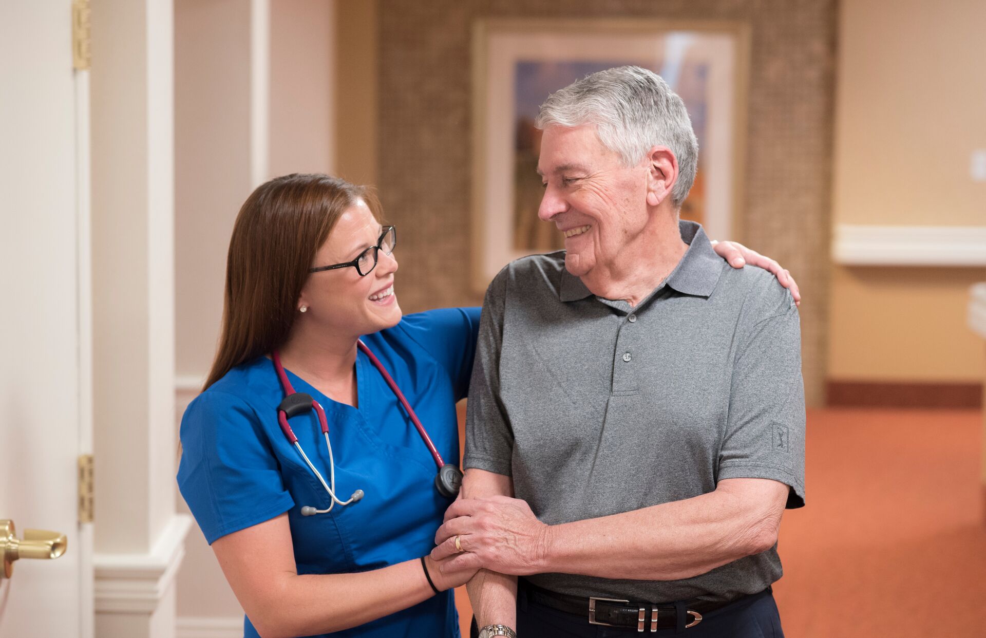 A senior man walks with assistance from a smiling nurse.